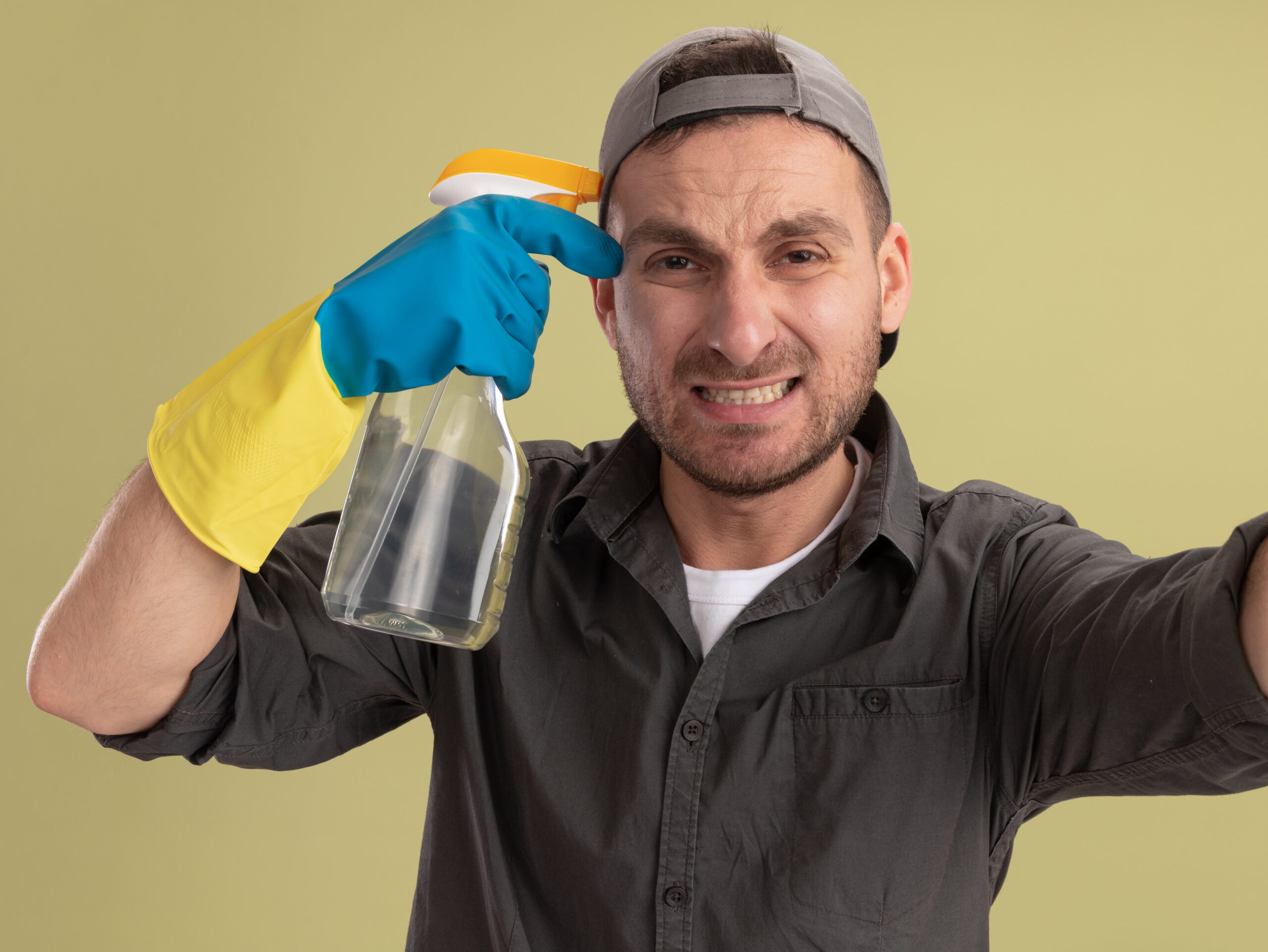 upset young cleaning man wearing casual clothes cap rubber gloves holding spray bottle looking crying standing green wall