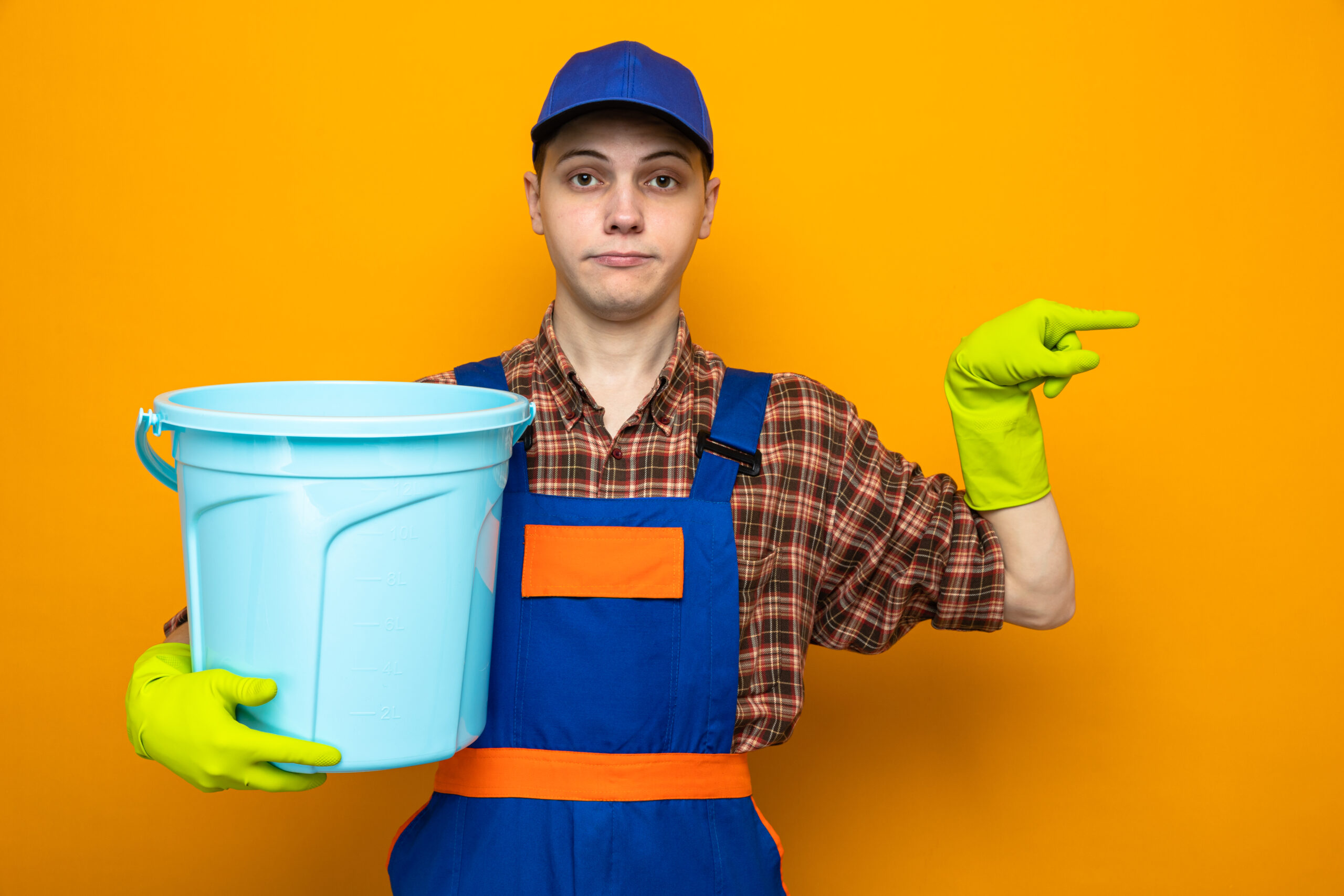 pleased points at side young cleaning guy wearing uniform and cap with gloves holding bucket isolated on orange background with copy space